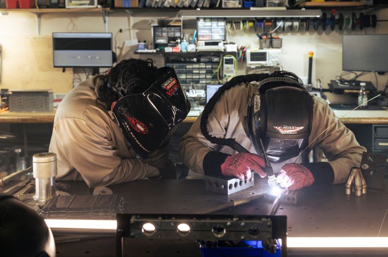 Two individuals welding at a workstation in a lab at KSU, surrounded by tools and equipment.