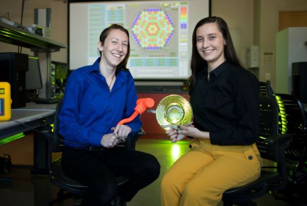 Two students sitting in a lab setting, smiling at the camera; one holds an orange tool, and the other holds a yellow glass dish, with a colorful hexagonal pattern displayed on a screen in the background.