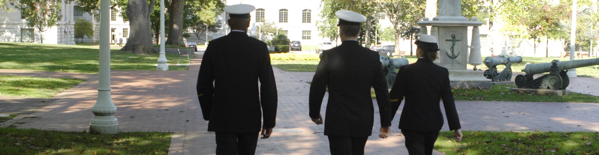 Three Navy ROTC members in uniform walking through a scenic campus with brick pathways, grassy areas, historical cannons, and buildings in the background.