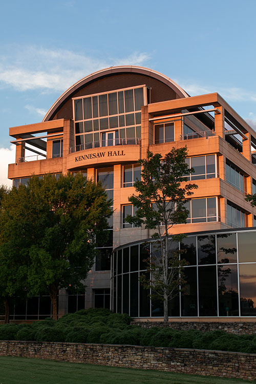 A warm evening light illuminates Kennesaw Hall, a modern building with a curved roofline and expansive glass windows. Trees and landscaping frame the structure.