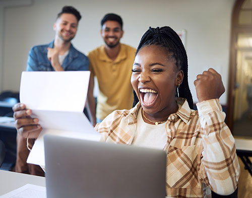 A young woman sits in front of a laptop, holding a piece of paper and celebrating a staff scholarship with a joyful expression and raised fist.