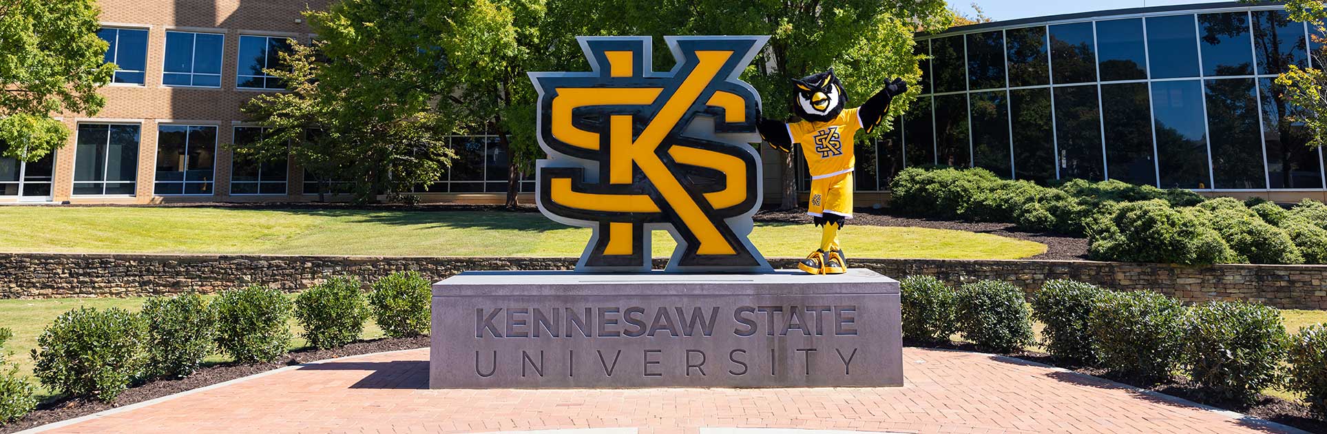 The Kennesaw State University mascot, Scrappy the Owl, stands next to the large KSU logo on a stone base. The logo and mascot are surrounded by green landscaping and a brick pathway.